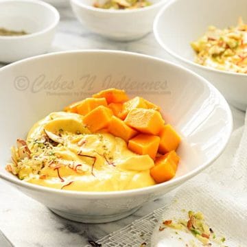 Close up shot of mango shrikhand in white bowl, nuts, cubed mangoes in bowls at the back.