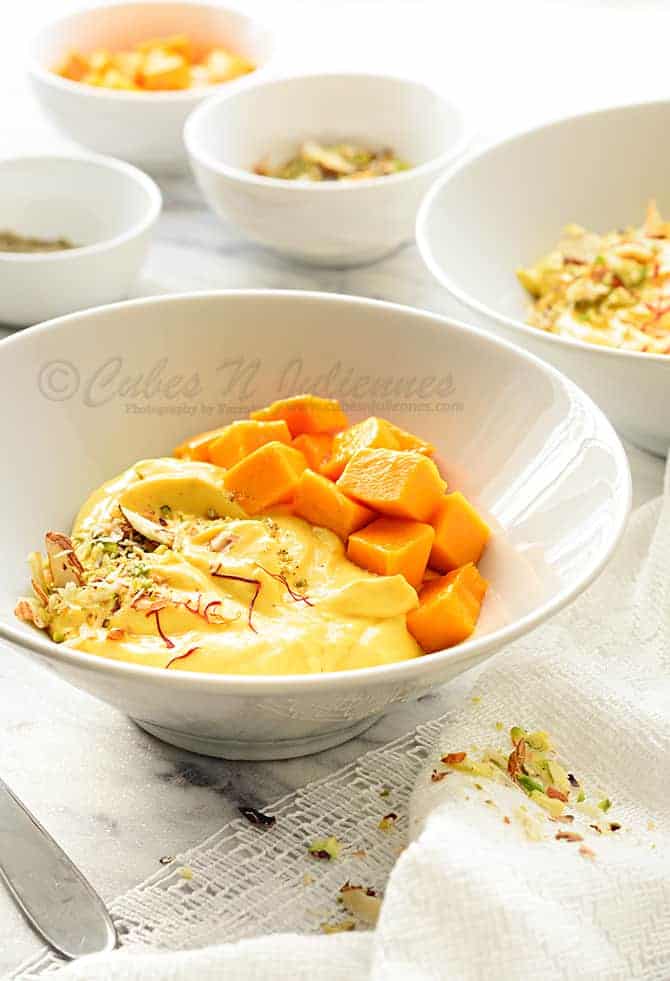 Close up shot of mango shrikhand in white bowl, nuts, cubed mangoes in bowls at the back.