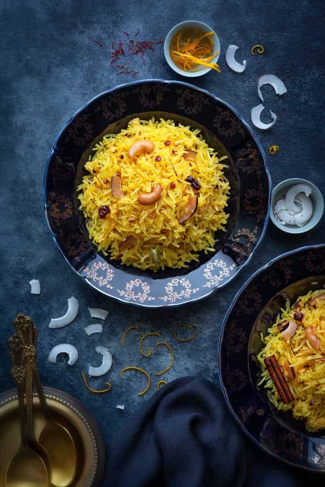 An over head shot of zarda served in served in two blue plates, coconut slices and orange zest in a small bowl and around