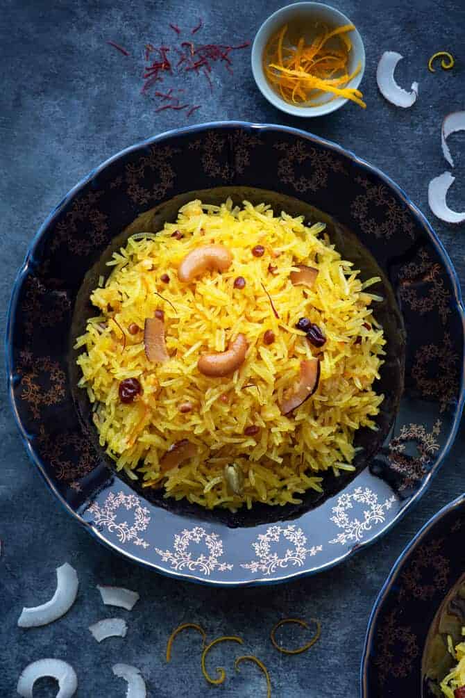 A close-up shot of Zarda pulao (sweet rice) served on a blue plate. Some orange zest, coconut slices spread around