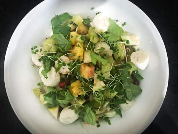 Mixed fruit chaat topped with mint, micro greens, spices and honey in white bowl.