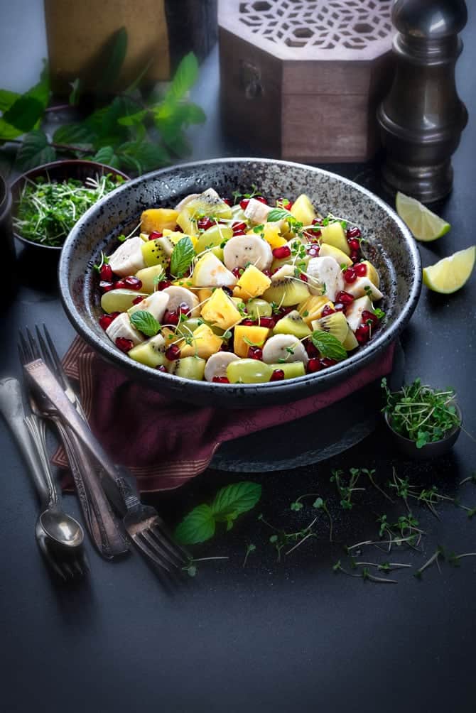 Spiced mixed fruit chaat in large black bowl, few spoons and fork on the side, few greens spread around.