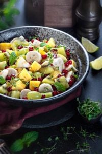 Close-up shot of fruit chaat salad in a large black bowl, lemon wedges on side.