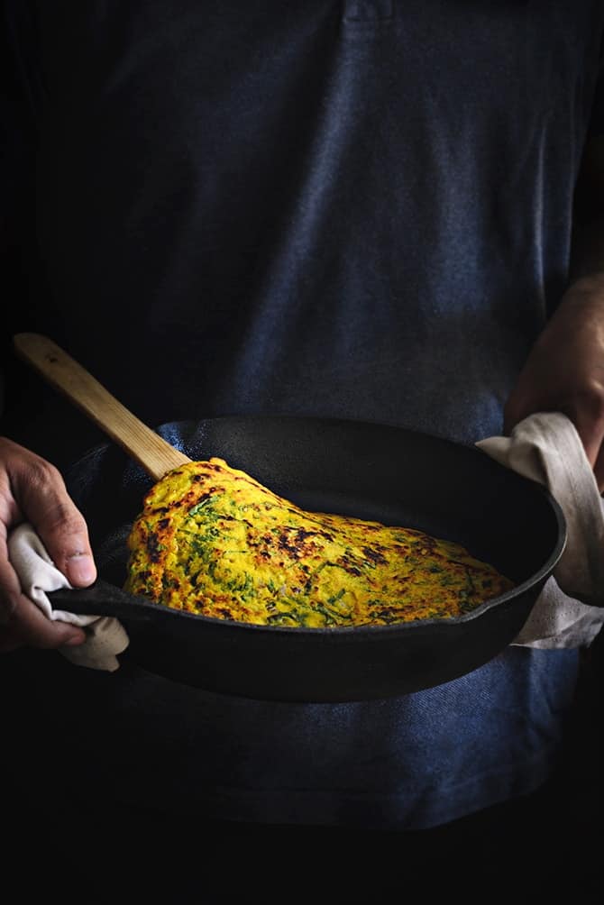 A pair of hands holding an iron pan with moong dal chilla in it.