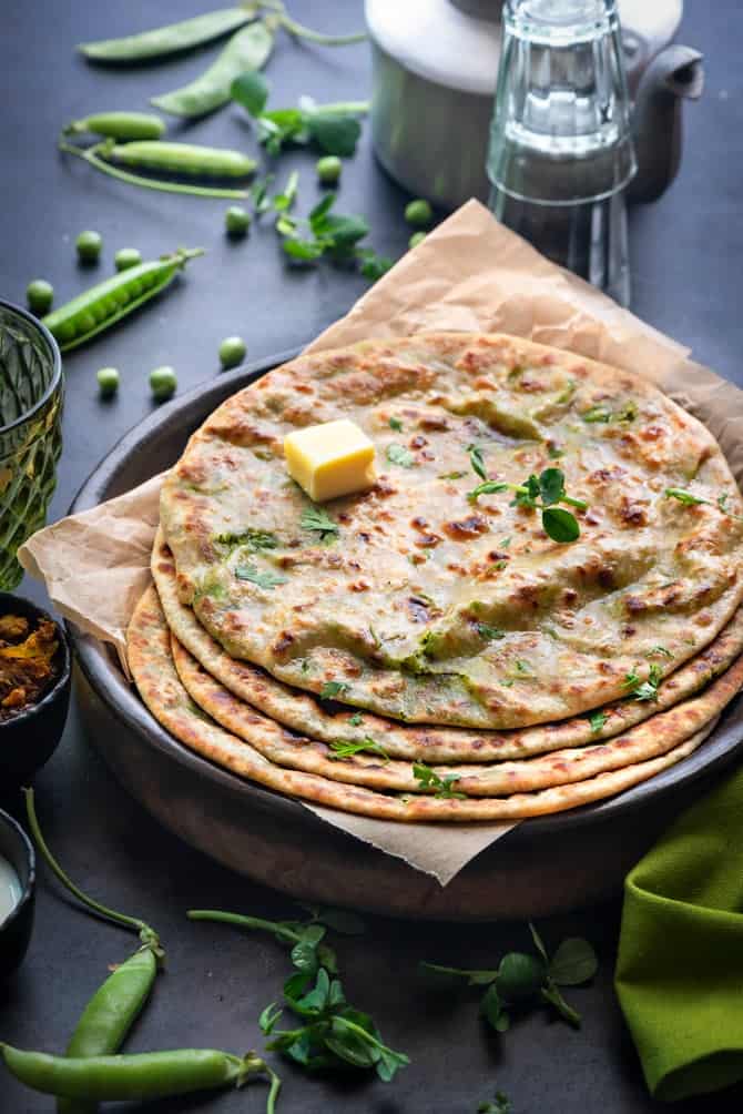 Stacked peas paratha or matar paratha topped with butter served on black plate. Teapot and cups at the back.