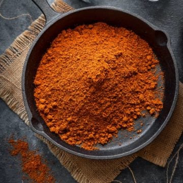 Homemade achari masala powder in cast iron pan with glass jars on the side.