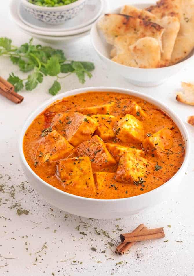 Creamy paneer butter masala gravy in white bowl with naan bread at the back.