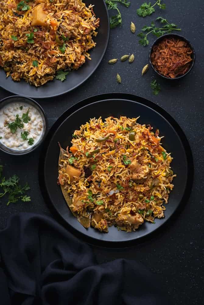 Instant pot vegetable biryani served in 2 black plates with Indian raita on side.