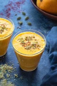 Close shot of mango lassi in two glasses, nuts, cardamom and saffron spread around.