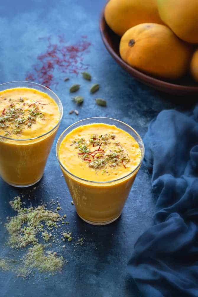 Mango Lassi served in two glasses, mangoes at the back, nuts, saffron and cardamom spread around.