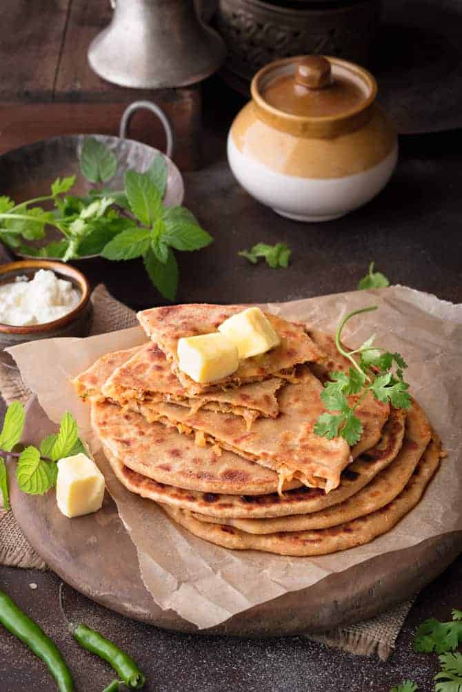 Stack of mooli paratha (radish paratha) topped with butter cubes, pickle jar and yogurt at the back.