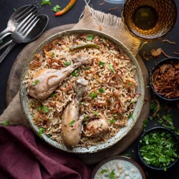 Spicy chicken pulao served in grey ceramic bowl, yogurt raita and fried onions on side.