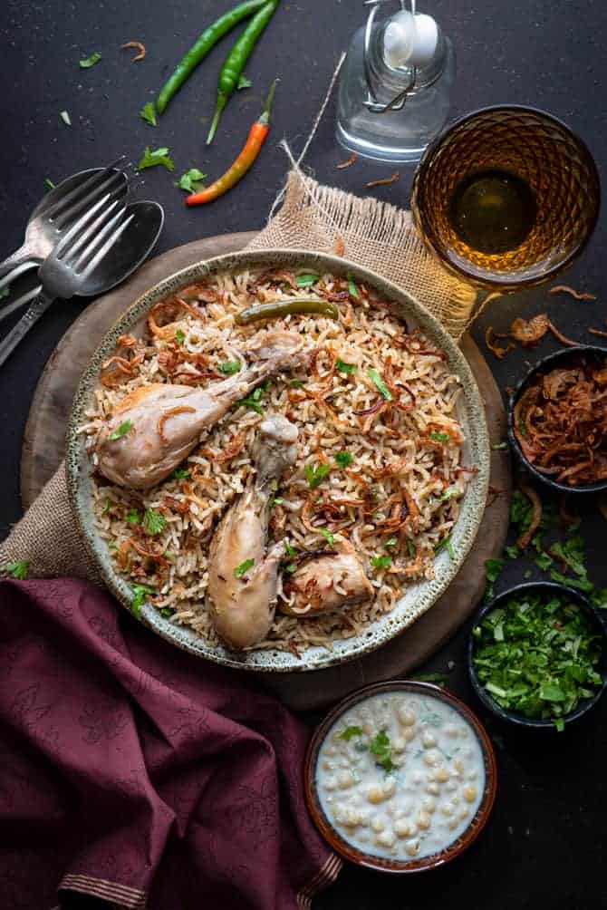 Spicy chicken pulao served in grey ceramic bowl, yogurt raita and fried onions on side.