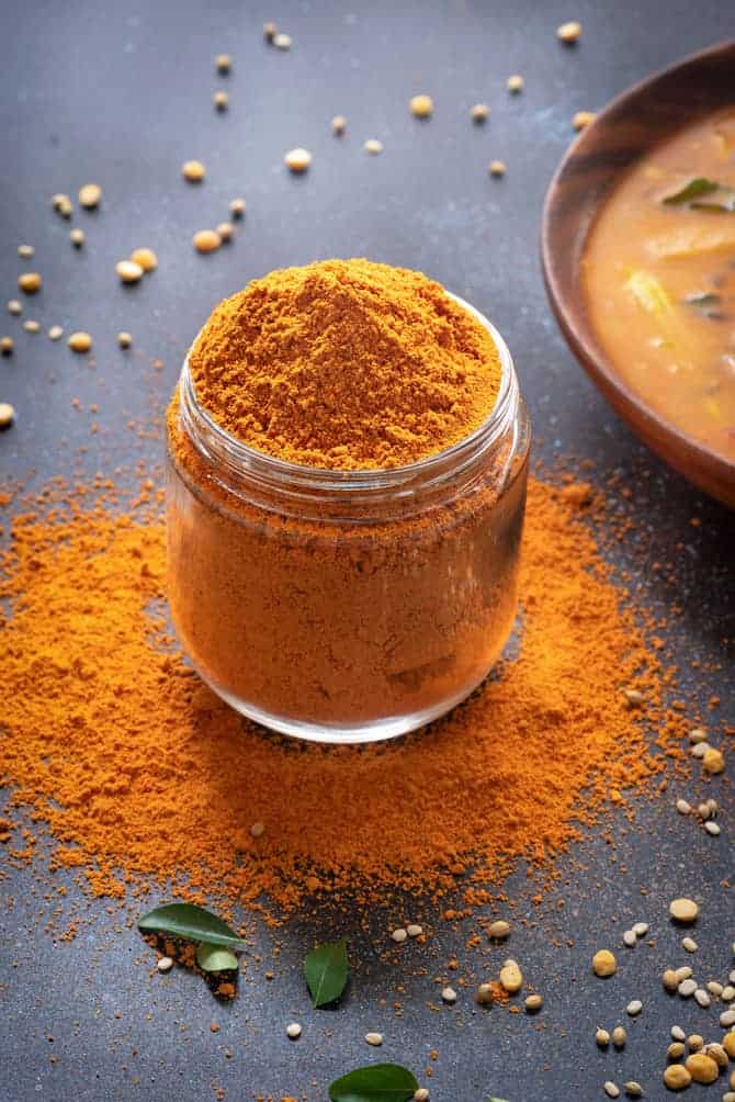 Sambar Masala powder in a glass jar, few curry leaves around, sambar in a bowl at the back.