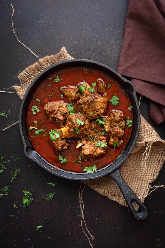 Overhead shot of Indian mutton curry or lamb curry in cast iron pan, some coriander leaves spread around.