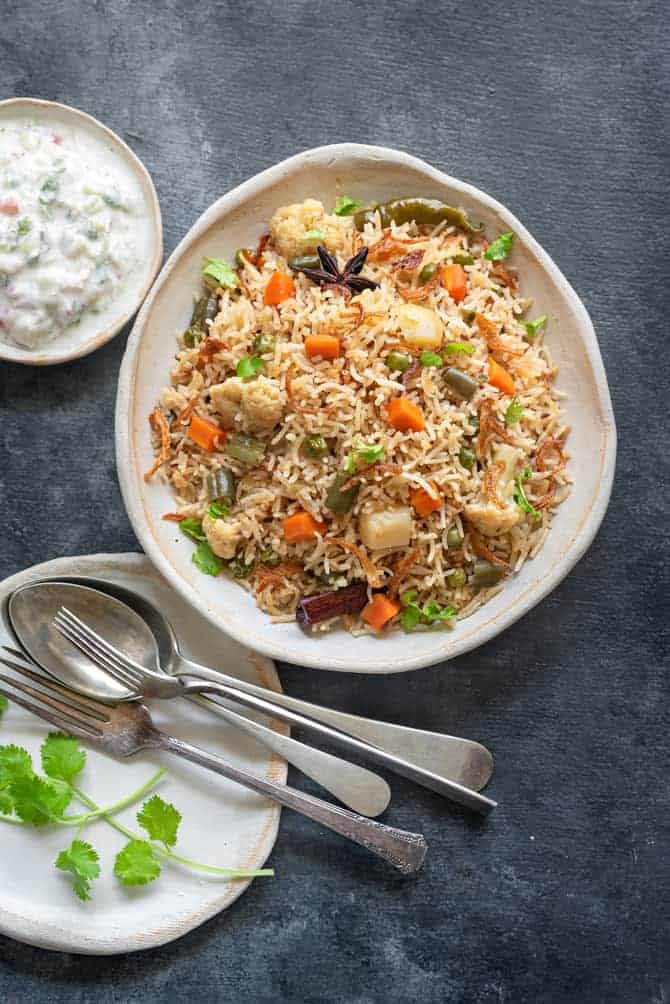 Vegetable pulao on white ceramic plate with raita and spoons on the side.