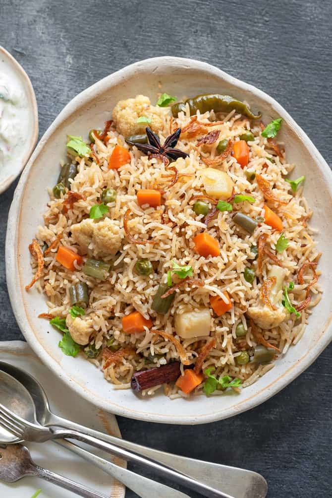 Close up shot of mix veg pulao on white ceramic plate, spoons and fork on the side.
