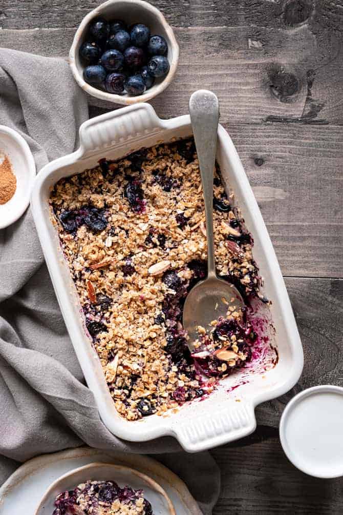Baked blueberry crisp in rectangular ceramic dish with a spoon, cream and some blueberries on side.