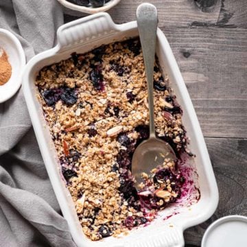 Blueberry crisp in rectangular ceramic dish with a spoon, cream and some blueberries on side.