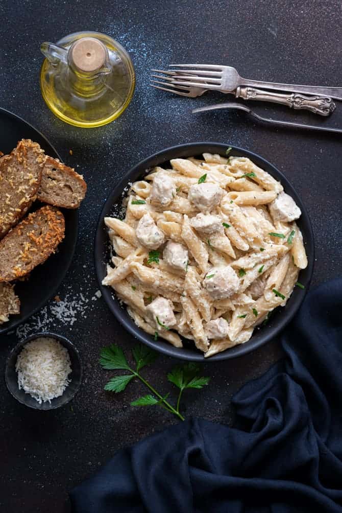 Overhead shot of chicken Alfredo pasta served in black plate, garlic bread and cheese on side.