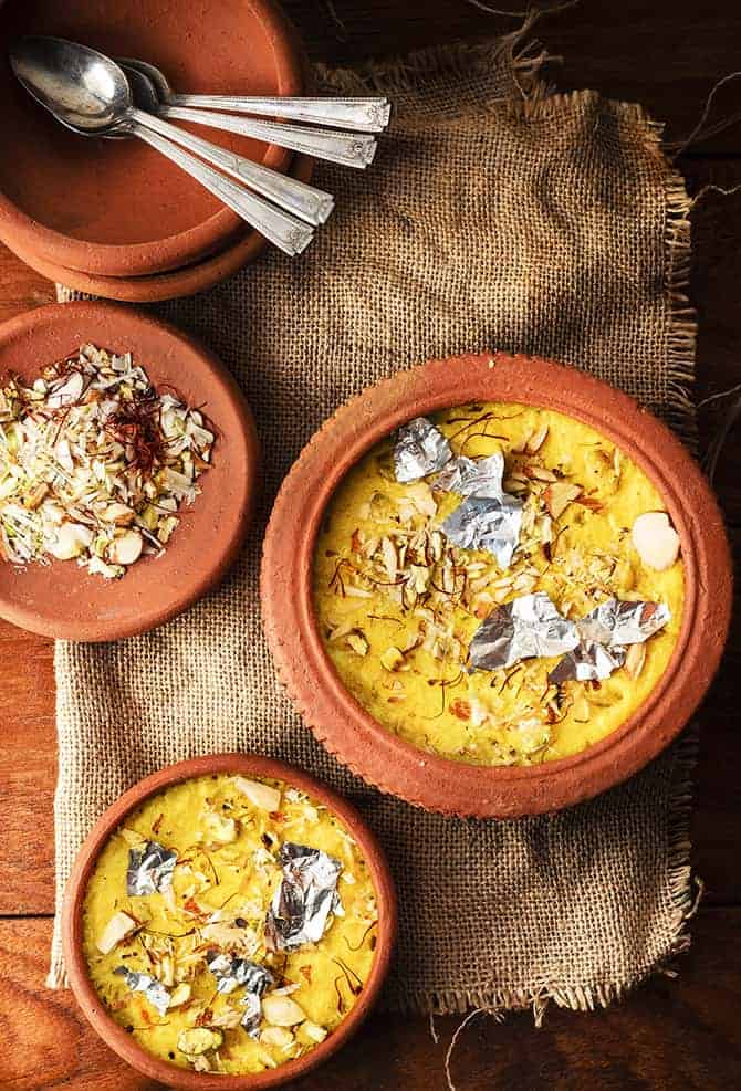 Overhead shot of kesar phirni sweet in two earthen pots, chopped dry fruits and spoon on side.