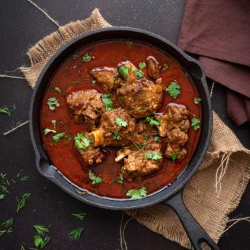 Overhead shot of Indian mutton curry or lamb curry in cast iron pan, some coriander leaves spread around.