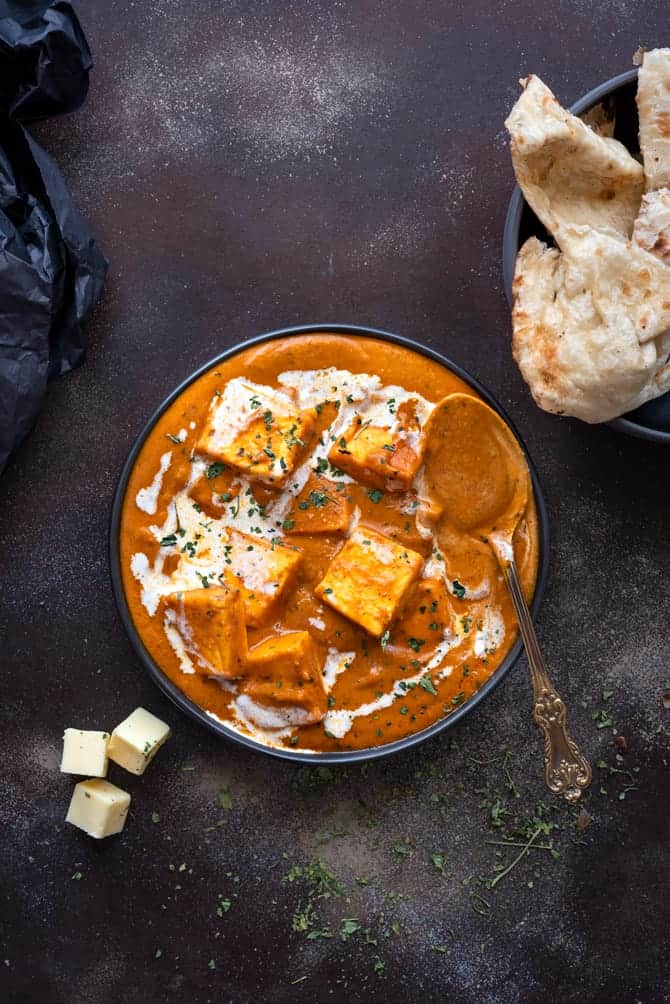 Overhead shot of paneer butter masala served in black plate with some naan bread on the side.