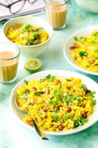 Close up shot of onion poha on a green plate and in bowl at the back, tea in glass on the side.