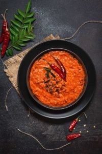 Kerala Style red coconut chutney on black plate with some red chilies and curry leaves spread around.