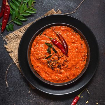 Kerala Style red coconut chutney on black plate with some red chilies and curry leaves spread around.