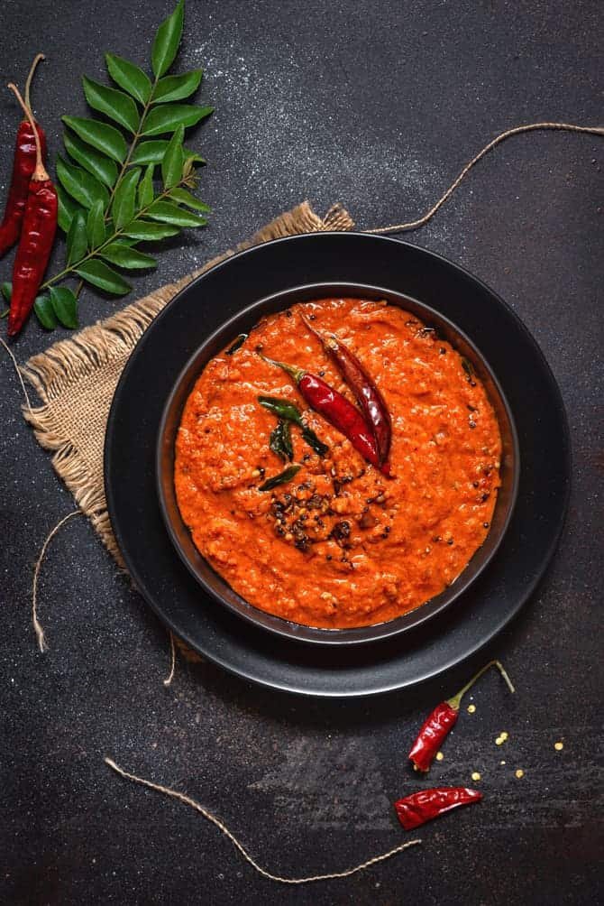 Kerala Style red coconut chutney on black plate with some red chilies and curry leaves spread around.