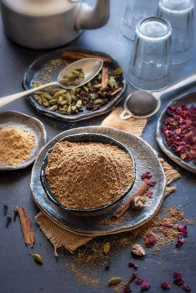 Chai masala powder in black bowl with some spice and rose petals spread around, glasses and teapot at the back.