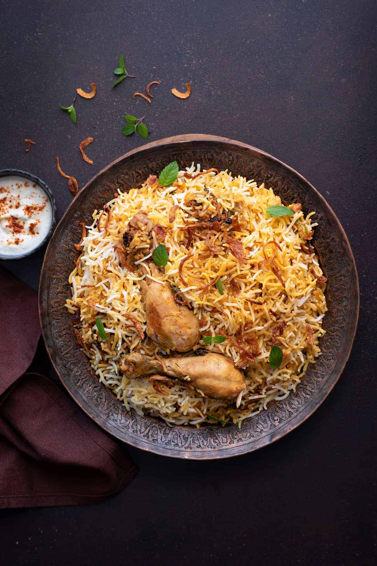 Overhead shot of chicken biryani served on copper plate with yogurt raita on the side.