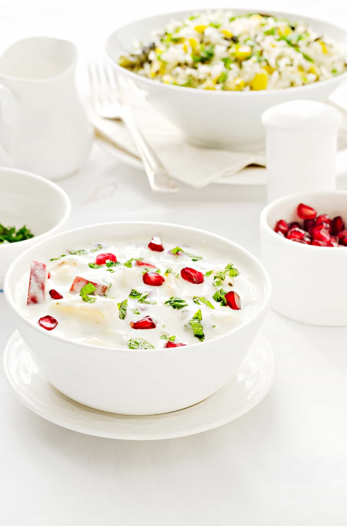 Mix Fruit Raita served in white bowl, rice pilaf and pomegranate in bowl at the back.