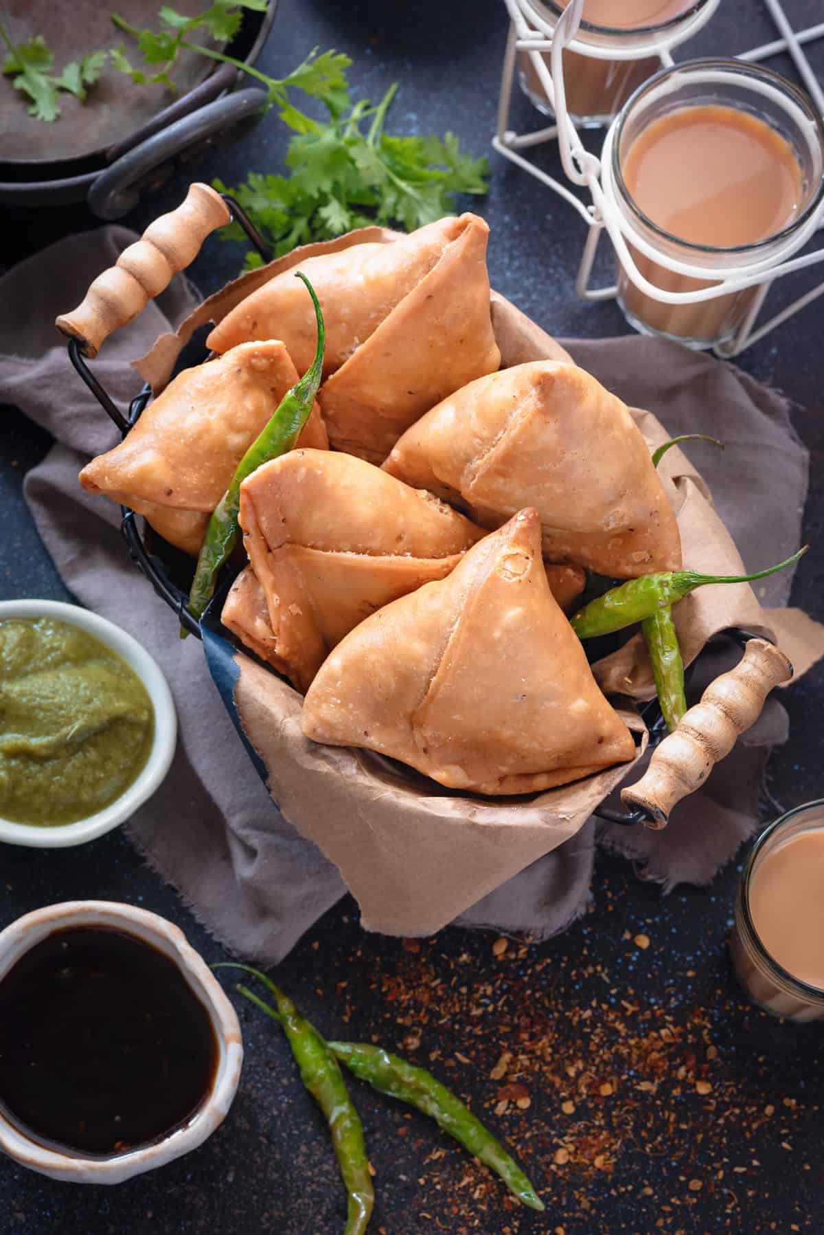 Overhead shot of flaky Indian Punjabi samosa in basket, tea and chutney on the side.