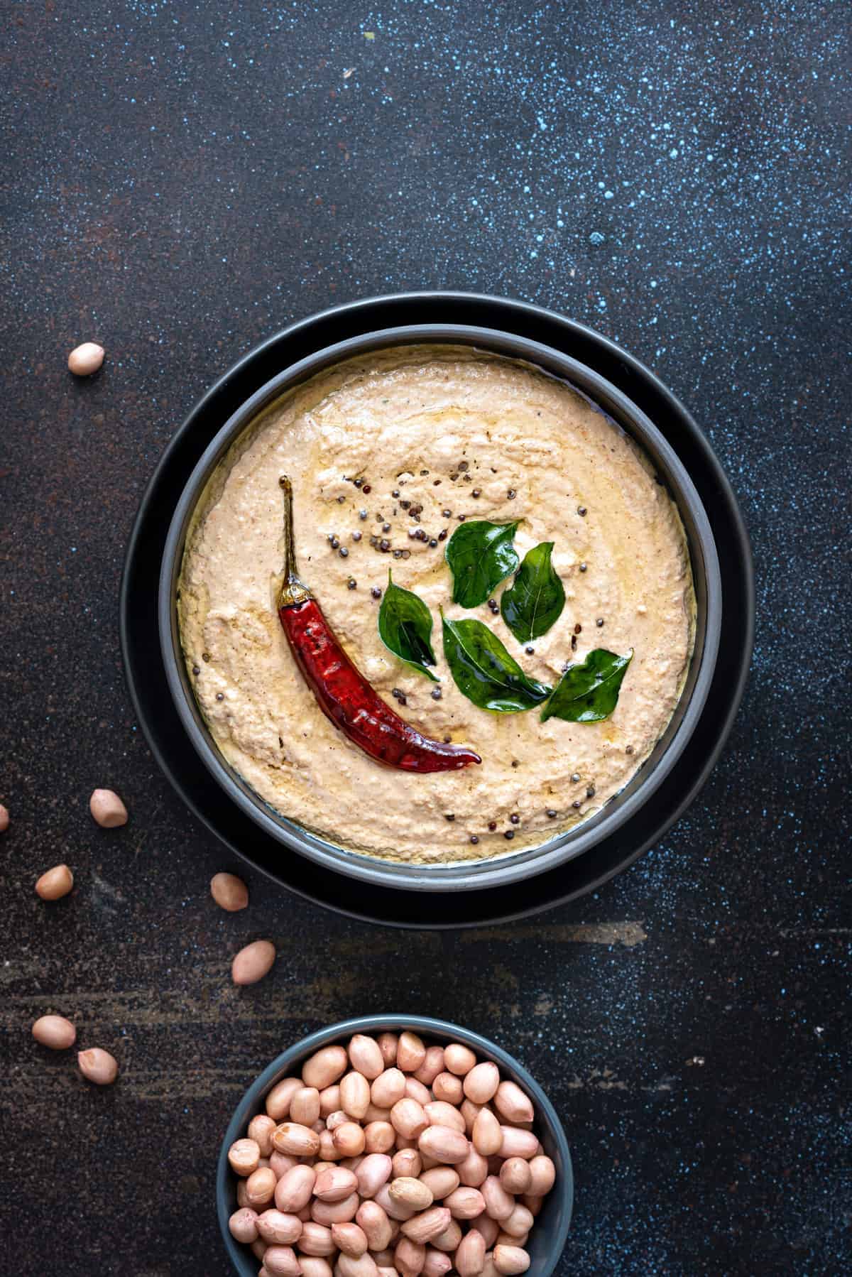 Overhead shot of peanut chutney in black bowl with few groundnut seeds spread around.