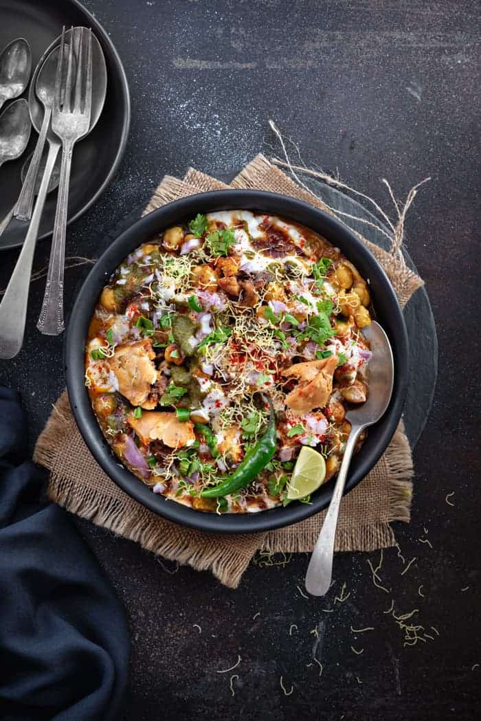 Overhead shot of chole samosa chaat served in black plate with a spoon into it.