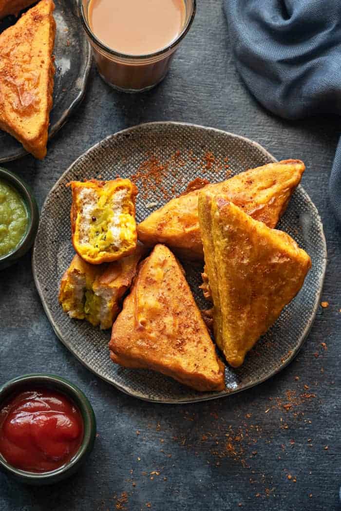 Stuffed bread pakora on two plates, one pakora cut open, chutney, sauce and tea on the side.