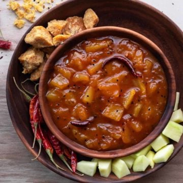 Aam ki launji in wooden bowl with jaggery, red chillies and raw mangoes spread around.