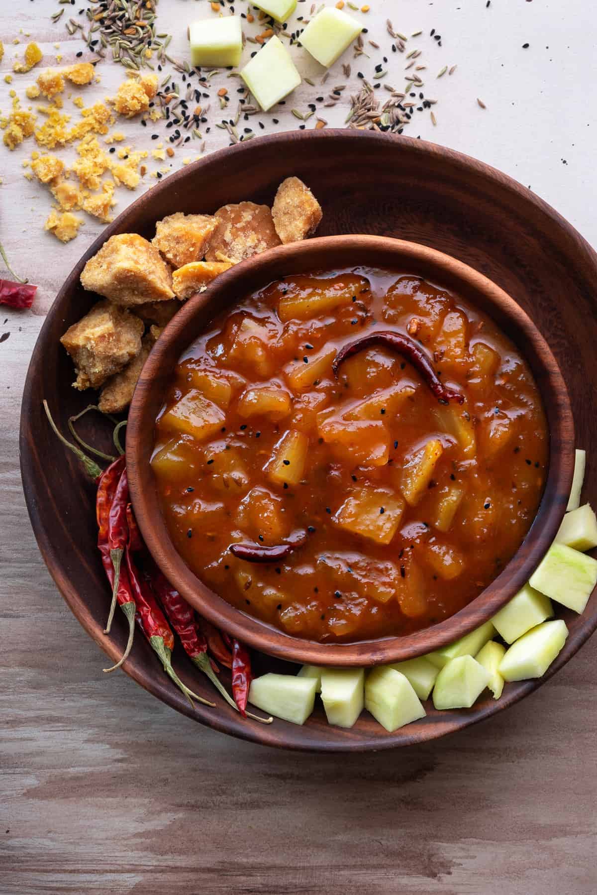 Aam ki launji in wooden bowl with jaggery, red chillies and raw mangoes spread around.