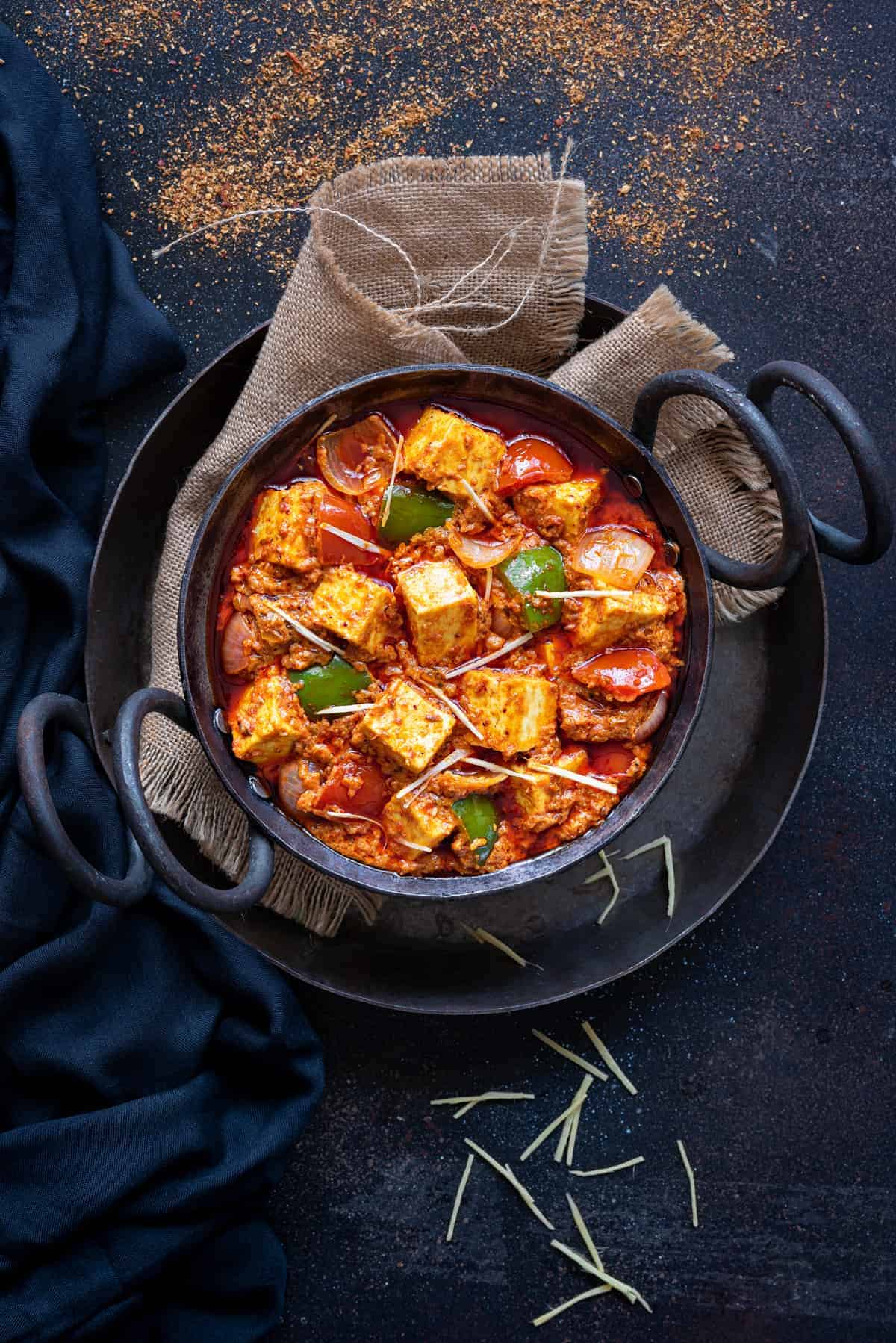 Kadhai paneer served in small black iron karahi, blue napkin and a piece of jute spread around.
