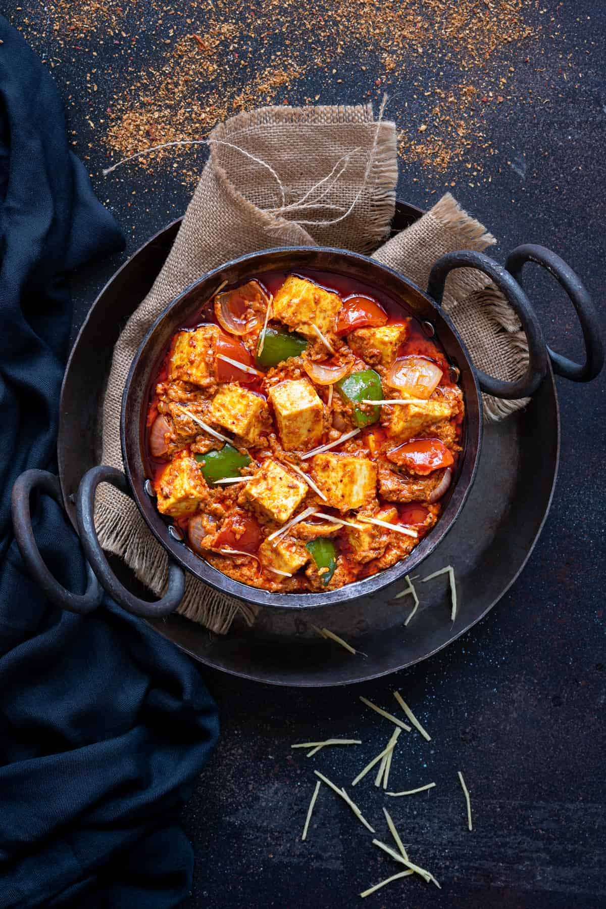 Kadhai paneer served in small black iron karahi, blue napkin and jute spread around.