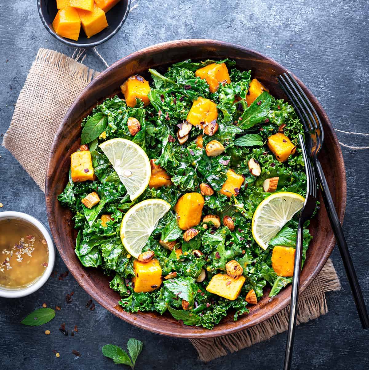 Mango Kale Salad in wooden bowl with 2 forks in it, cut mangoes and dressing in two small bowls on side.