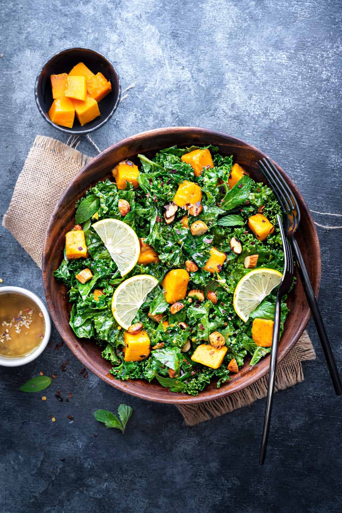 Mango Kale Salad in wooden bowl with forks, mango cubes and dressing in two small bowls.