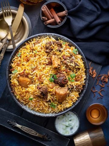 Overhead shot of Bombay Mutton Biryani served in large black bowl with raita on the side.