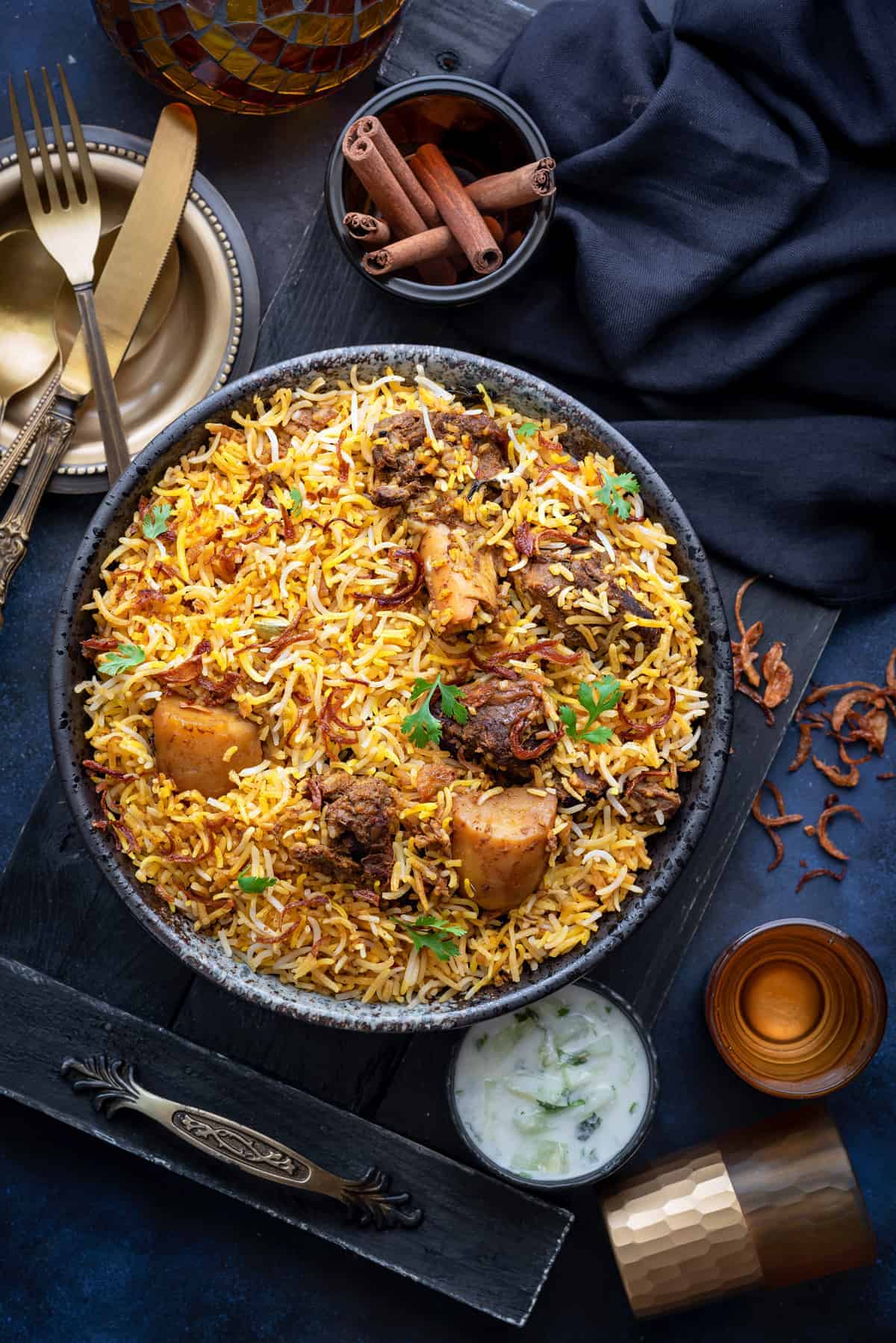 Overhead shot of Bombay Mutton Biryani served in large black bowl with raita on the side.