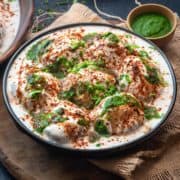 Dahi Vada topped with sweet and spicy chutney and served in black plate, green chutney at the back.