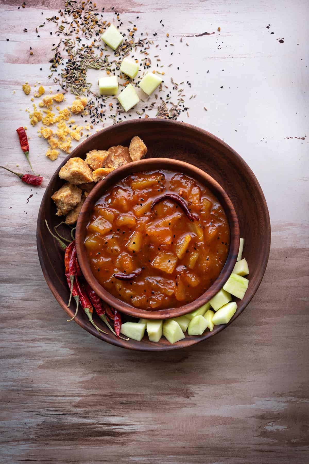 Sweet raw mango chutney in wooden bowl with jaggery, mango and red chillies spread around.