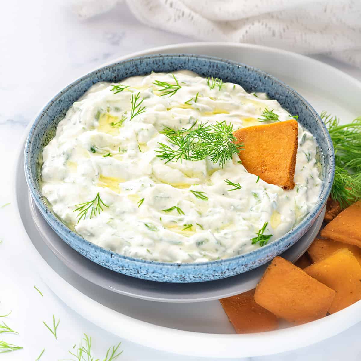Authentic Greek Tzatziki sauce served in blue bowl with some pita chips on the side.