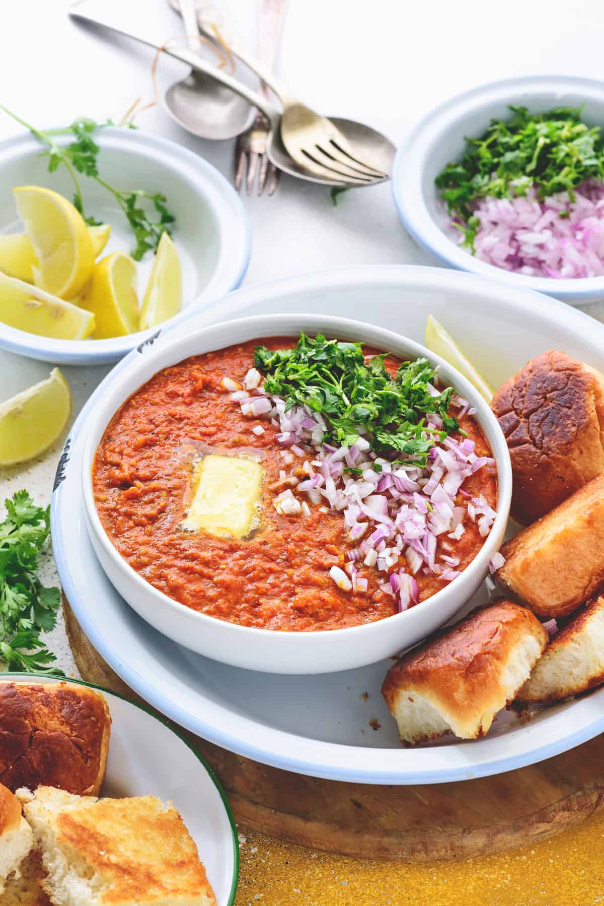 Garnished Pav Bhaaji served in white bowl with buttered buns, chopped onions and lemon wedges.